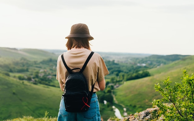 Detrás de una chica turista se encuentra en una montaña con un hermoso paisaje verde Mujer hipster en ropa casual y con una mochila se encuentra en el borde de la montaña