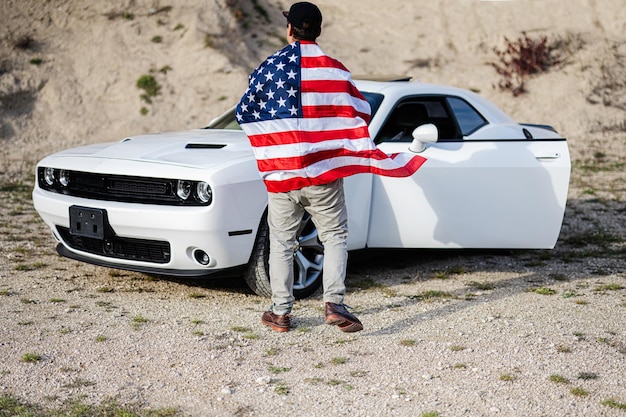 Detrás del hombre con la bandera de Estados Unidos cerca de su muscle car blanco americano en carrera