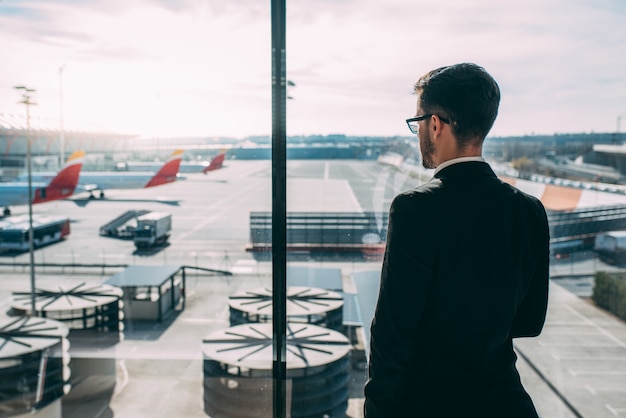 Detrás de un joven hombre de negocios de pie con la maleta en el aeropuerto esperando el vuelo