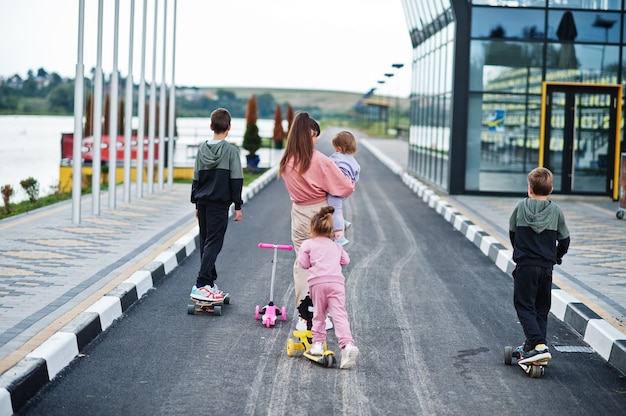 Detrás de la joven madre elegante con cuatro hijos al aire libre. La familia deportiva pasa el tiempo libre al aire libre con patinetes y patines.