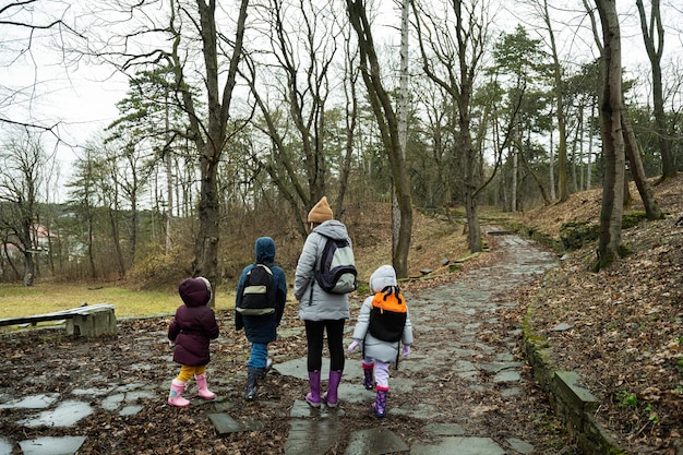 Detrás de mamá e hijos con mochilas caminando juntos por el camino de piedra del bosque después de la lluvia