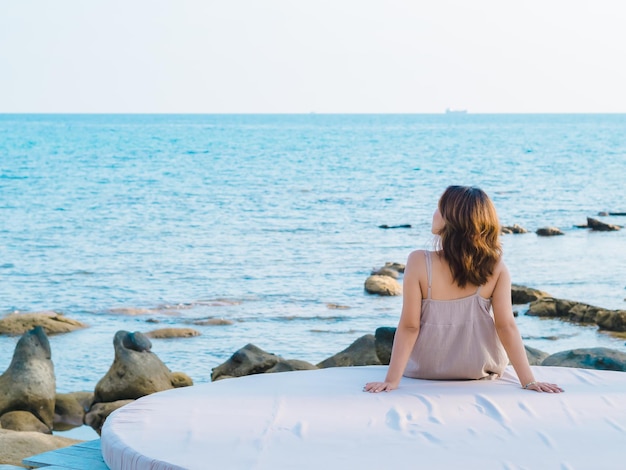 Detrás de una mujer asiática vestida de forma casual sentada en un reposabrazos en un cojín frente a la playa Seascape en vibraciones de verano Una mujer solitaria relajando la sonrisa en el mar y mirando lejos Vacaciones de viaje de vacaciones