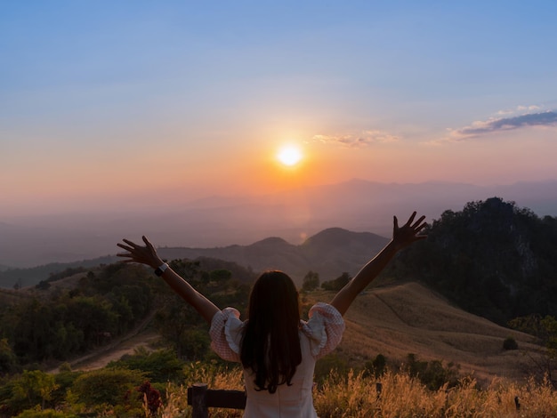 Detrás de una turista asiática levanta las manos en la cima de la colina con una hermosa vista a la montaña al atardecer en el mirador de Doi Samer Dao en Nan, Tailandia.