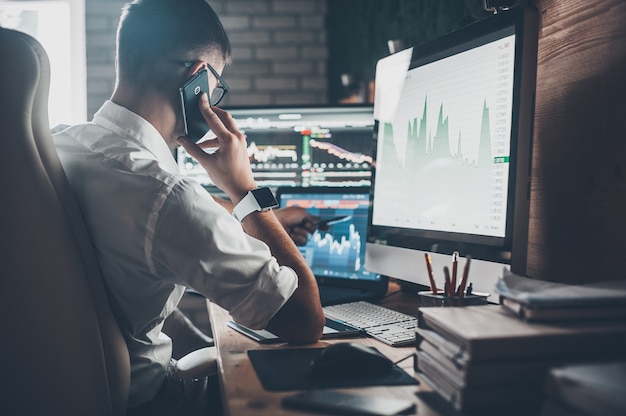 Dia agitado de trabalho no trabalho. Vista traseira de um jovem em trajes casuais falando ao telefone e trabalhando enquanto está sentado à mesa no escritório criativo