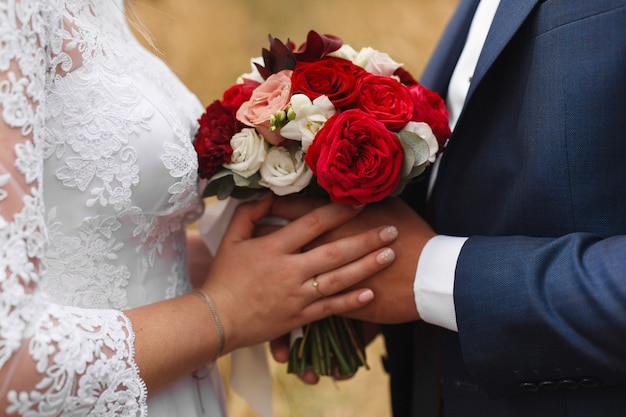 Día de la boda al aire libre. Momento romántico de boda. Retrato de novios cogidos de la mano suavemente. recién casados con ramo de novia rojo de cerca