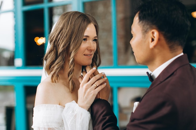 Día de la boda Hermosa novia europea y su novio asiático posando contra el telón de fondo de cafés y grandes ventanales