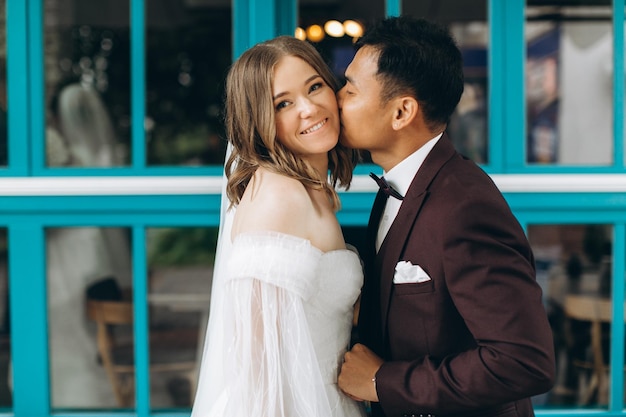 Día de la boda Hermosa novia europea y su novio asiático posando contra el telón de fondo de cafés y grandes ventanales