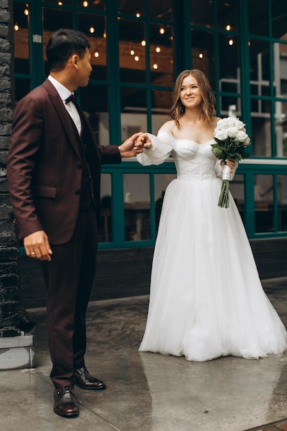 Día de la boda Hermosa novia europea y su novio asiático posando contra el telón de fondo de cafés y grandes ventanales