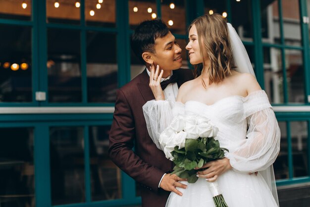 Día de la boda Hermosa novia europea y su novio asiático posando contra el telón de fondo de cafés y grandes ventanales