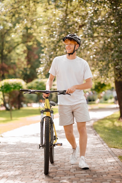 Dia bom. ruivo sorridente de capacete e roupas esportivas com bicicleta andando no parque em dia ensolarado
