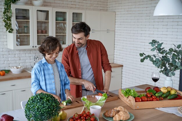 Dia da família, jovem pai e filho preparando uma salada e cortando vegetais frescos em pé no