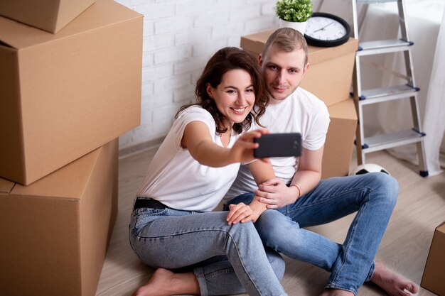Dia da mudança - retrato de um jovem casal sorridente tirando uma foto de selfie com smartphone, cercado de caixas de papelão em uma nova casa ou apartamento