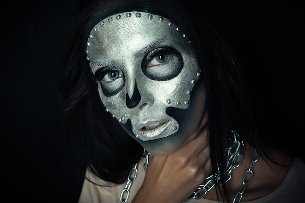 Dia das bruxas, feriados, estilo de vida, pessoas, beleza, conceito criativo - tema de Halloween e maquiagem criativa: modelo de menina bonita com corpo preto com máscara prateada pintura do crânio em fundo escuro no estúdio