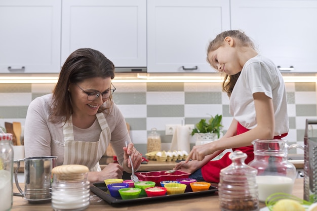 Dia das mães, mãe e filha criança de 9, 10 anos preparando cupcakes juntas em casa na cozinha, mulher ensinando criança a cozinhar