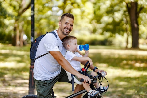 Dia de folga para a família Beber água e andar de bicicleta Menino senta-se em uma cesta de bicicleta e bebe água de uma garrafa azul enquanto um homem adulto olha diretamente para a câmera e senta-se em uma bicicleta atrás de uma criança