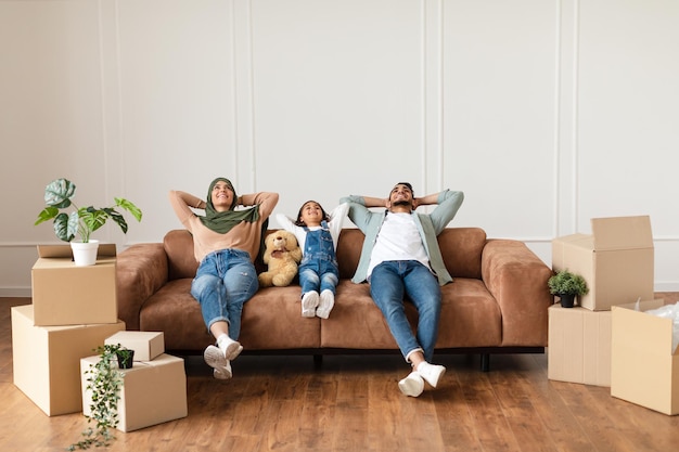 Foto dia de mudança. linda família árabe relaxando no sofá em uma nova casa com caixas de papelão ao redor. homem feliz, mulher com lenço na cabeça e menina deitada no sofá olhando para cima, cópia gratuita