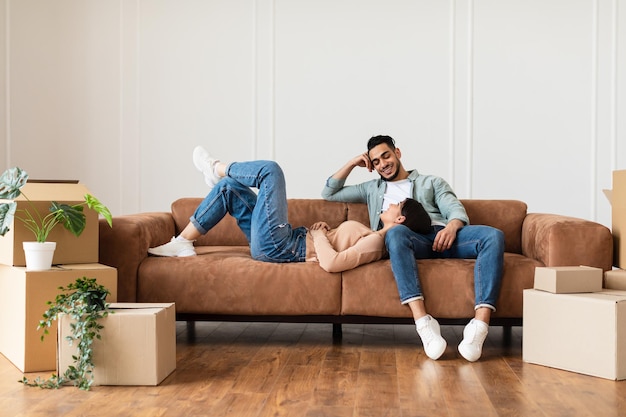 Dia de mudança. Lindo casal relaxando no sofá em uma nova casa com caixas de papelão ao redor. Homem feliz e mulher deitada no sofá e sonhando, discutindo o futuro design de interiores, espaço de cópia gratuita