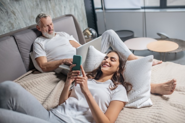 Dia de sorte. Mulher sorridente e alegre de cabelos escuros com smartphone e marido grisalho concentrado com laptop na cama