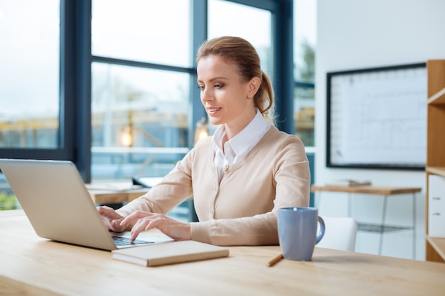 Dia de trabalho. Mulher de negócios agradável concentrada sentada à mesa enquanto usa um laptop e prepara um relatório importante