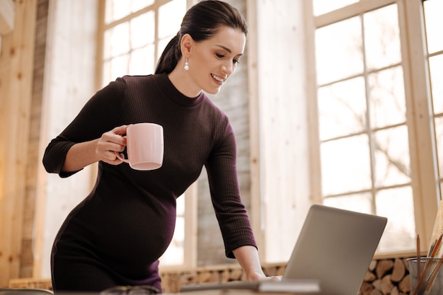 Dia de trabalho. mulher de negócios grávida e entusiasmada bebendo chá enquanto digita no laptop e fica