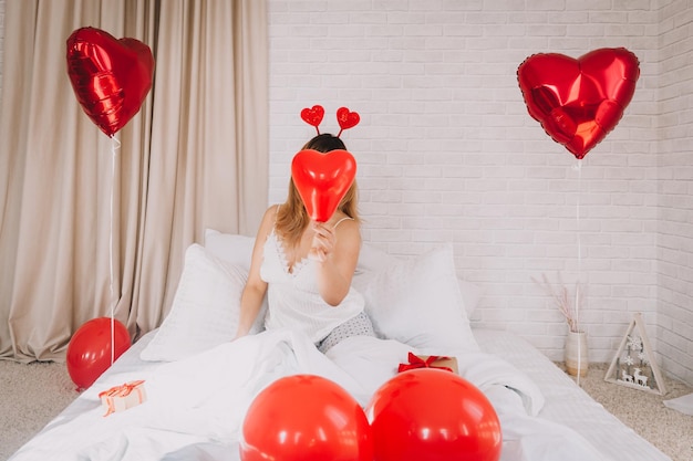 Dia dos Namorados, Dia da Mulher. Jovem garota caucasiana sentada na cama e comemorando o dia dos namorados segurando um balão em forma de coração nas mãos