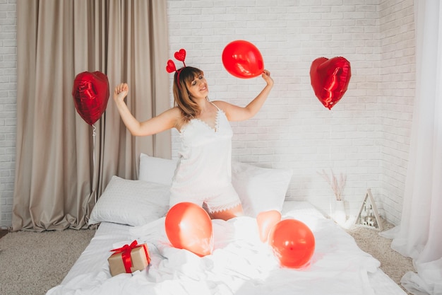 Dia dos Namorados, Dia da Mulher. Jovem garota caucasiana sentada na cama e comemorando o dia dos namorados segurando um balão em forma de coração nas mãos