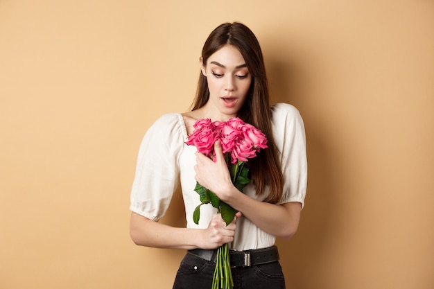 Dia dos Namorados. Menina surpresa recebe um presente do amante no encontro, parecendo espantada com o lindo buquê de flores, segurando rosas sobre fundo bege.