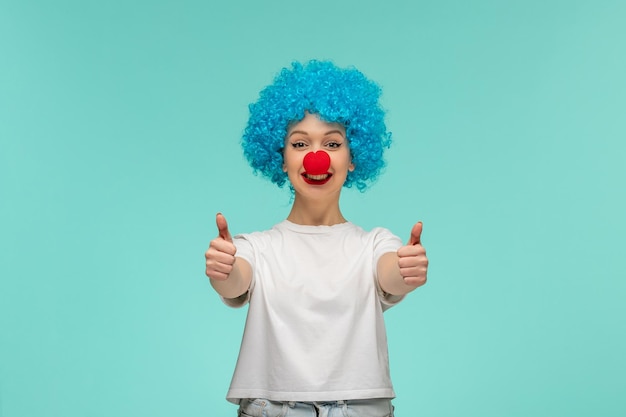 Dia dos tolos de abril, garota animada, gesto de mãos grandes sorrindo com nariz vermelho em cabelo azul de fantasia de palhaço