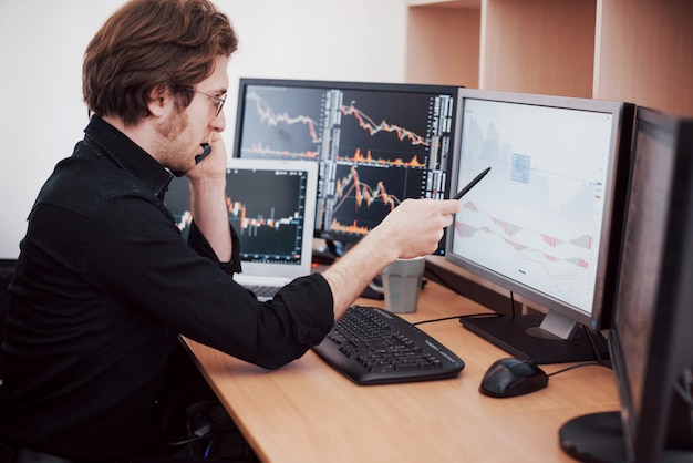 Dia estressante no escritório. Jovem empresário de mãos dadas no rosto enquanto está sentado na mesa do escritório criativo. Conceito do gráfico da finança dos estrangeiros de troca da bolsa de valores