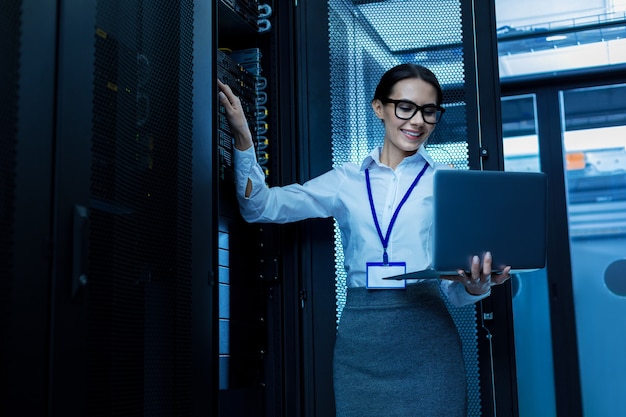 Día increíble. Feliz hermosa mujer trabajando en un gabinete de servidor y sosteniendo su computadora portátil