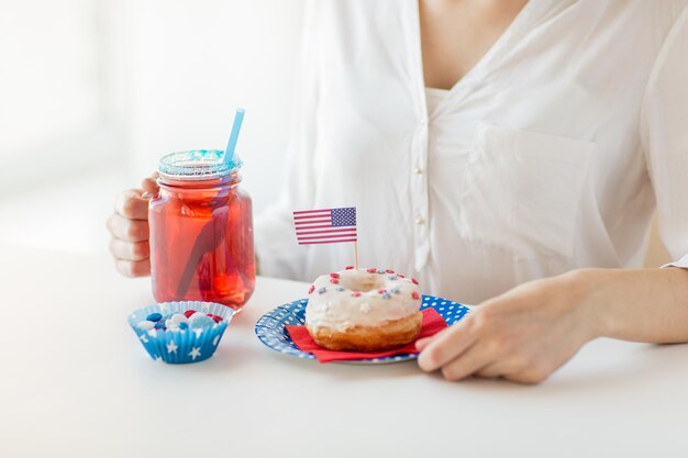 día de la independencia, celebración, patriotismo y concepto de vacaciones - cerca de una mujer comiendo donut dulce glaseado, bebiendo jugo de un gran tarro o taza de vidrio y celebrando el 4 de julio en la fiesta de casa