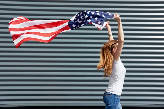 Día de la independencia y concepto patriótico. Chica activa con cabello largo y jengibre corriendo con ondeado por el viento USA flag