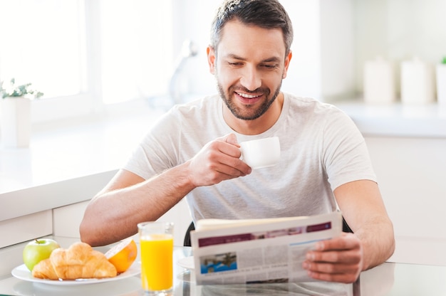 Día de inicio de buenas noticias. Apuesto joven leyendo el periódico y sonriendo mientras toma café y desayuna en la cocina