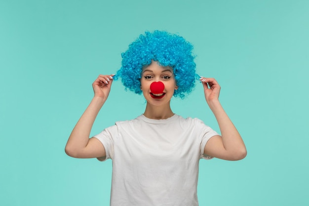 Foto día de los inocentes linda chica con nariz roja en un disfraz de payaso tirando de un mechón de cabello azul divertido