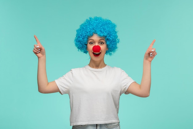 Día de los inocentes niña feliz dedos apuntando hacia arriba nariz roja en un disfraz de payaso pelo azul