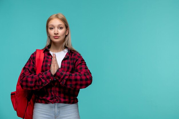 Día internacional de los estudiantes linda chica encantadora en camisa roja a cuadros rezando y vistiendo mochila roja