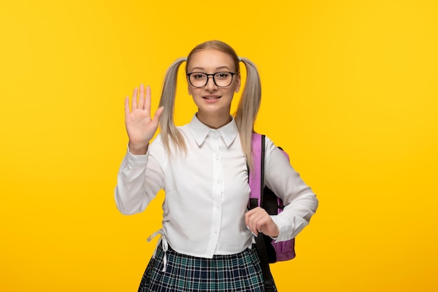 Día mundial del libro rubia joven colegiala diciendo hola gesto con la mano sonriendo con gafas y uniforme