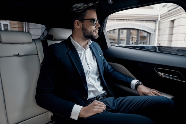 Día ocupado. Apuesto joven en traje completo mirando por la ventana mientras está sentado en el coche