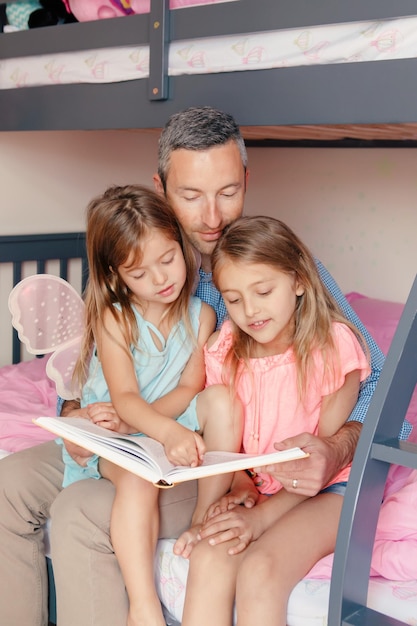 Foto día de los padres padre leyendo un libro a las hijas niñas familia de tres personas sentadas juntas leyendo bo