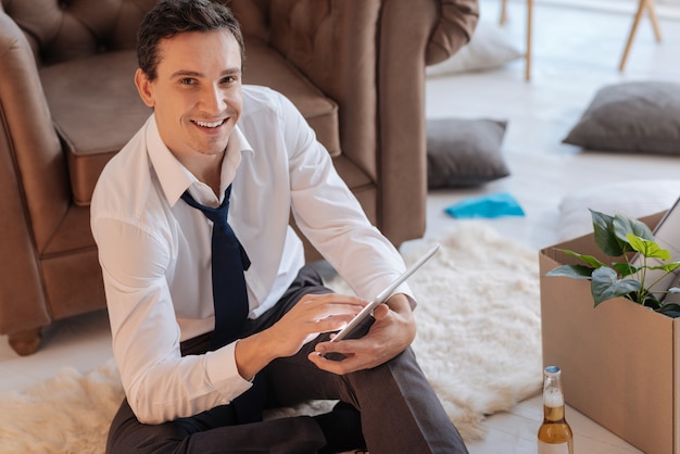 Día productivo. Hombre inteligente sonriente positivo sentado en el suelo y sosteniendo una tableta moderna mientras una botella de alcohol de pie a su lado