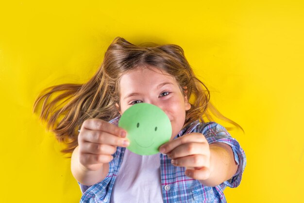 Día de la salud mental Proteger a los niños del concepto de estrés Terapia de depresión infantil de pensamiento positivo Linda niña preadolescente europea sostiene un emoticono sonriente de papel en su mano sobre fondo amarillo