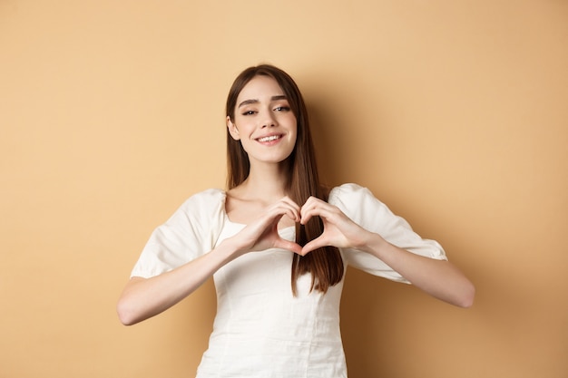 Día de San Valentín atractiva joven en vestido blanco sonriendo y mostrando gesto de corazón de pie en ...