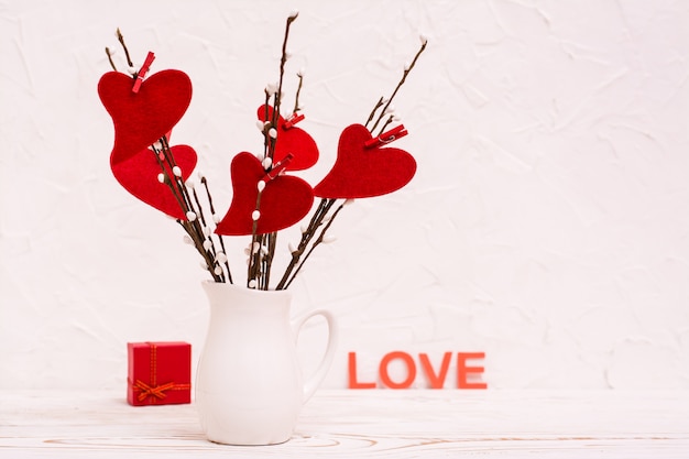 Día de San Valentín. Corazones rojos hechos de fieltro en ramas de sauce en una jarra blanca sobre la mesa, un regalo y la palabra "amor".