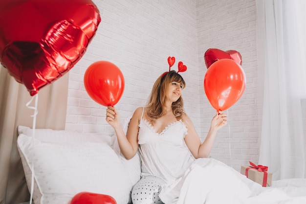 Día de San Valentín, Día de la Mujer. Joven caucásica sentada en la cama y celebrando el día de san valentín sosteniendo un globo en forma de corazón en sus manos