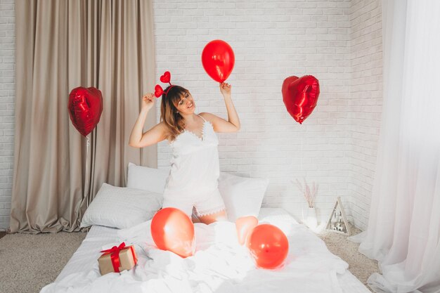 Día de San Valentín, Día de la Mujer. Joven caucásica sentada en la cama y celebrando el día de san valentín sosteniendo un globo en forma de corazón en sus manos