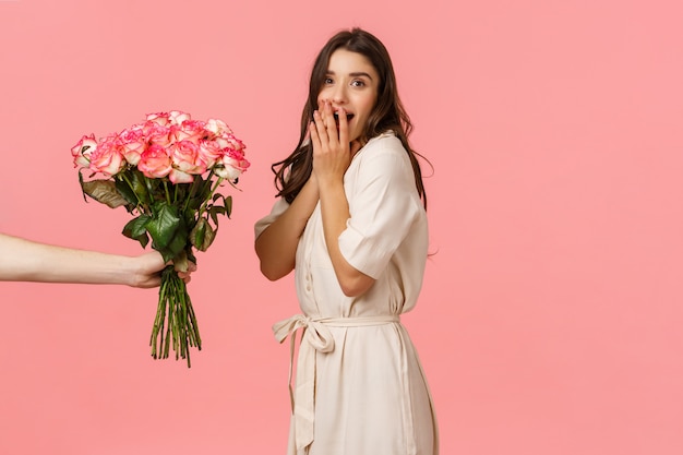 Día de San Valentín, entrega y concepto de belleza. Mujer sorprendida que jadea y se ve sorprendida cuando alguien le da un hermoso ramo de flores, felicitaciones con aniversario, pared rosa