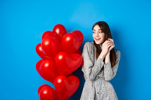 Día de San Valentín. Hermosa chica romántica soñando con una cita, de pie cerca de globos de corazón encantadores y sonriendo, fondo azul