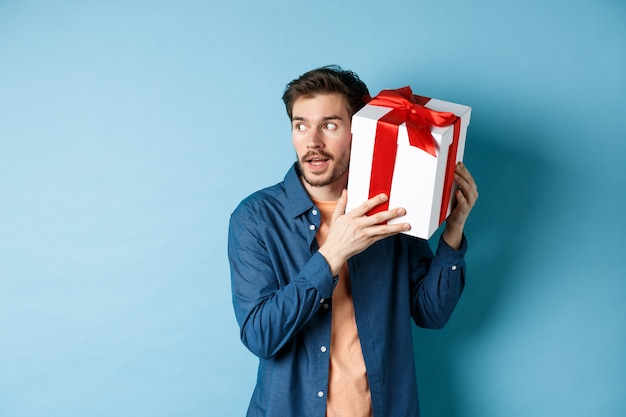 Día de San Valentín. Novio curioso con caja de regalo cerca de la oreja, tratando de adivinar qué hay dentro del presente sorpresa, de pie sobre fondo azul.