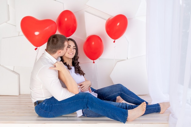 El día de San Valentín, una pareja enamorada sentada sobre un fondo claro abrazándose entre globos de corazones