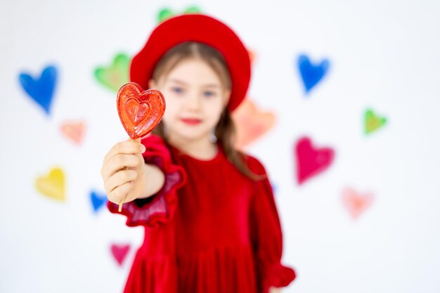 Foto día de san valentín una pequeña niña sonriente en una boina roja feliz sostiene o lamia una piruleta grande en forma de corazón y se regocija contra el fondo de corazones coloridos